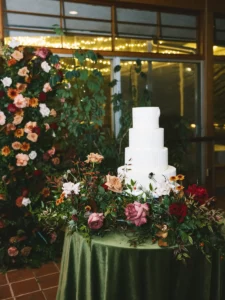 A three tiered white cake on a green velvet table linen surrounded by greenery and pink and purple roses