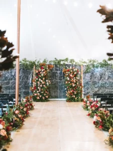 A walkway for an indoor ceremony lined with greenery and red and pink florals