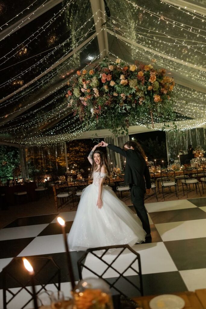 bride and groom on black and white dance floor under clear top wedding tent draped with twinkle lighting and large floral chandelier suspended in tent