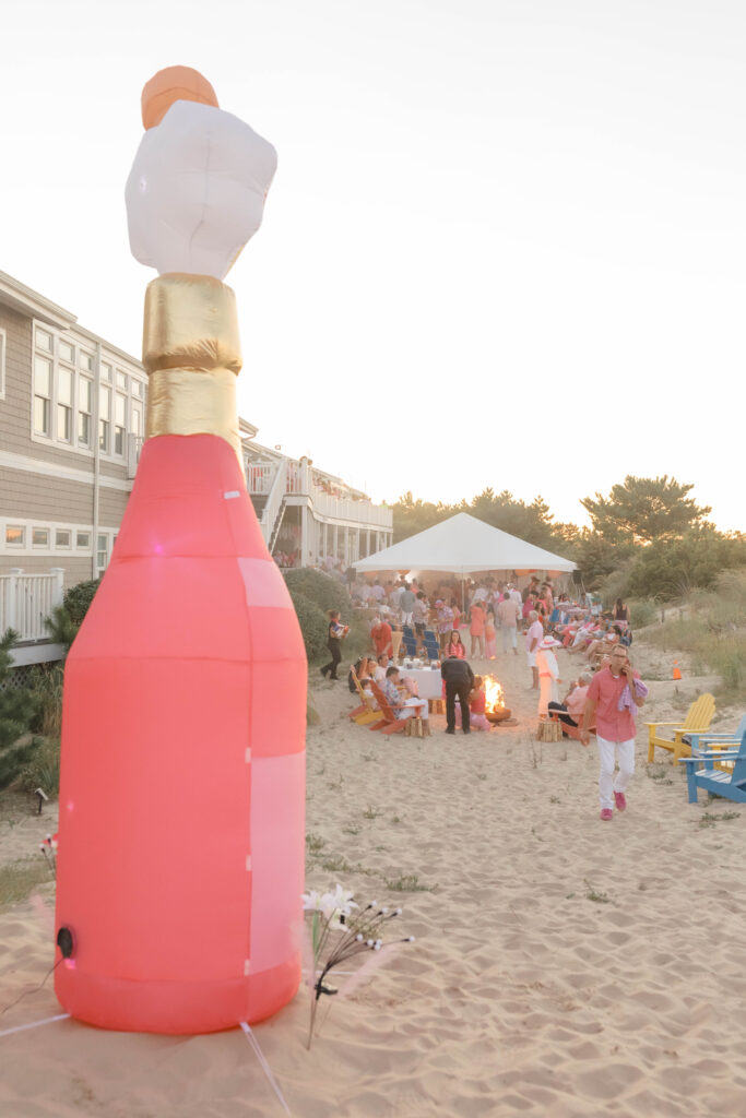 blow up pink champagne bottle set up at a beach party on sand with white tent in background