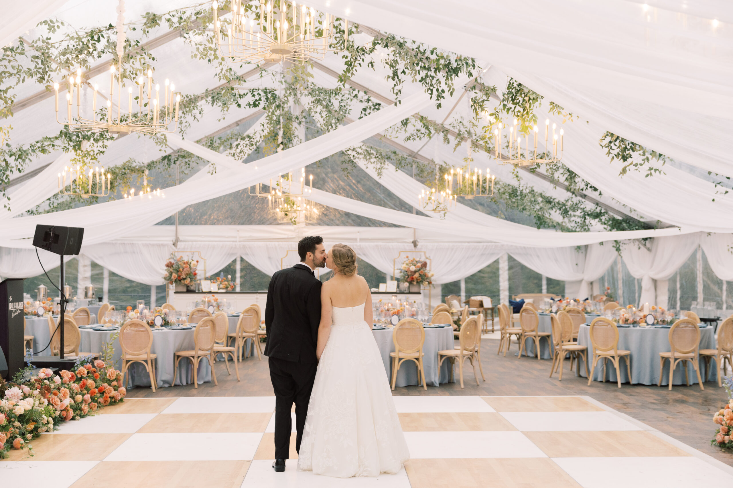 COCKTAIL-MULLER-BWP-121 bride and groom standing on dance floor under a clear top tent with fabric draping and greenery draped from ceiling